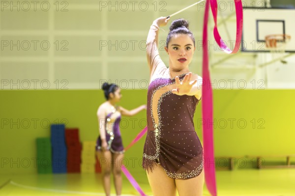 Young disabled rhythmic gymnast in elegant attire performs with a pink ribbon inside a sports hall.