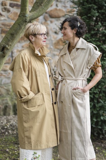 Two fashion women in stylish beige trench coats share a joyful moment outdoors. They stand near a