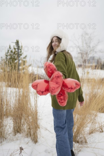 A woman wearing a green sweater and fur hat holds red heart shaped balloons in a snowy park. She