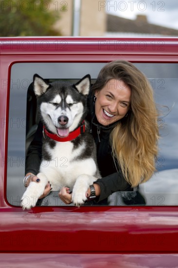 A joyful woman with long hair leans out of a red car window, holding a happy husky dog wearing a