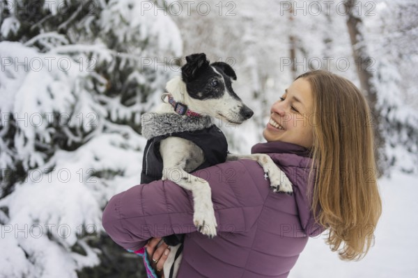 A woman in a purple jacket holds her dog lovingly in a snowy forest. Her smile and the snowy scene