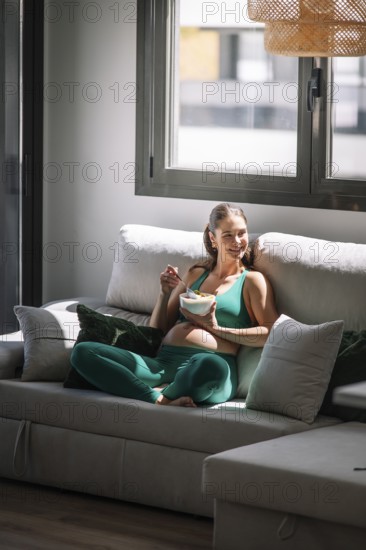 Pregnant woman in a green yoga outfit relaxes on a sofa, enjoying a healthy bowl of fruit. Sunlight