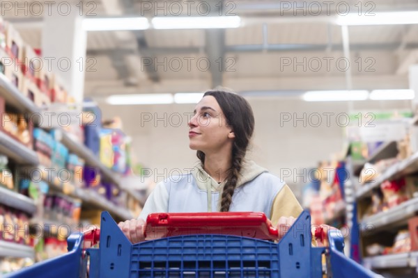 A woman with a braided ponytail glances upwards while pushing a red shopping cart in a supermarket