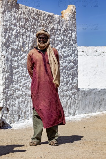 A man in traditional Tuareg clothing stands in front of a sunlit, plastered wall, Local people in
