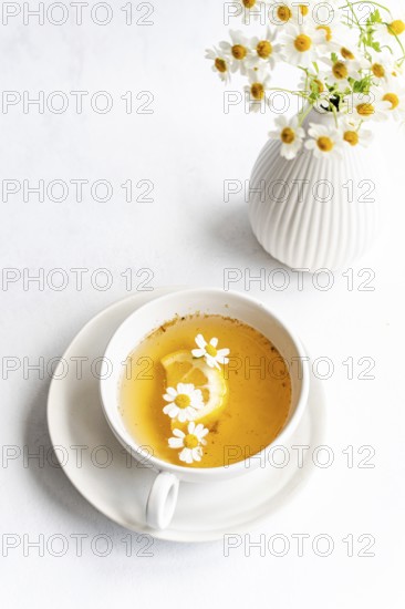 Top view of a white cup containing tea with daisies and lemon slice on a saucer. Nearby, a white