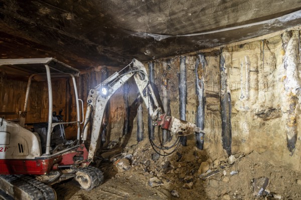 An excavator operates in an underground construction site, tearing through rock and earth. The dim