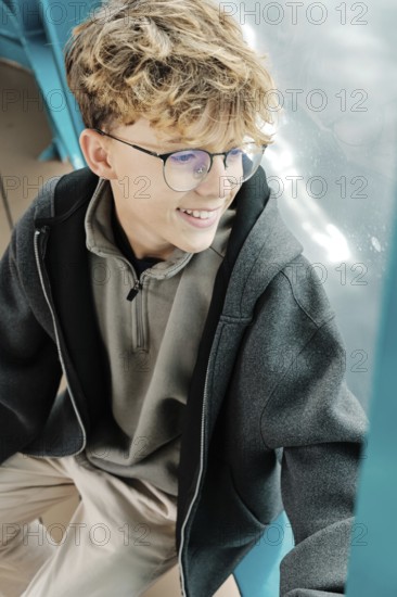 A curly-haired teenager with glasses sits by a window, smiling as he enjoys a ski resort in the