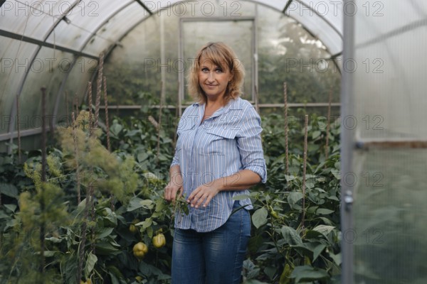 A woman in casual attire stands amidst lush plants inside a greenhouse. She tends to the greenery,