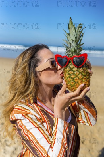 Woman playing with a pineapple wearing red heart sunglasses, standing on a sandy beach by the