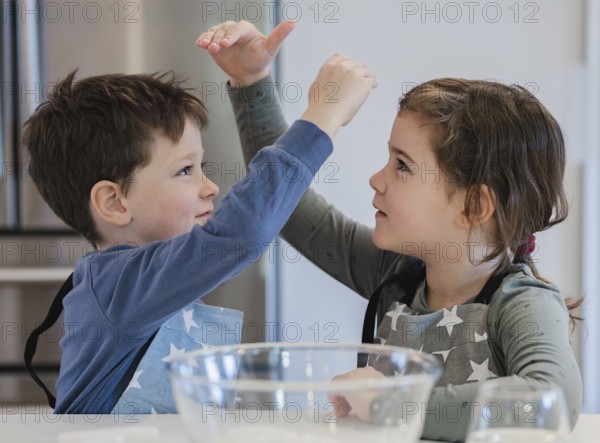 Two young siblings wearing star patterned aprons are joyfully interacting in a kitchen setting. A