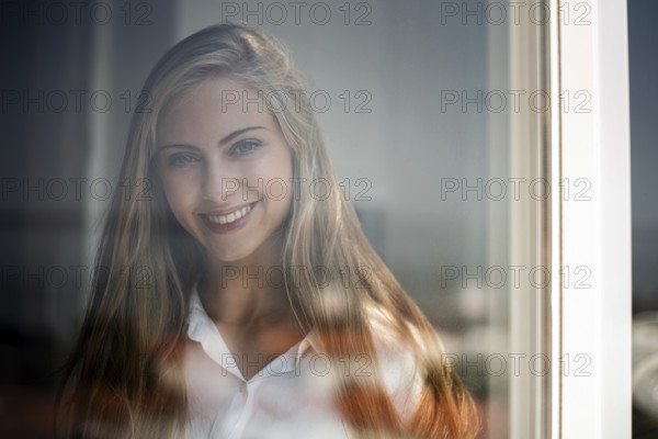 A young woman smiles gently through a glass window, captured in soft natural light. Her relaxed