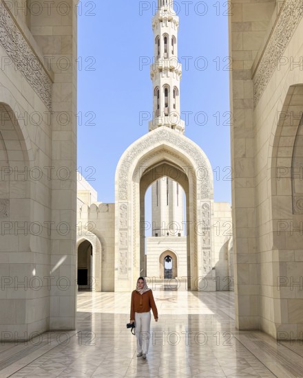 A woman wearing an headscarf walks through the grand architecture of the Sultan Qaboos Mosque in