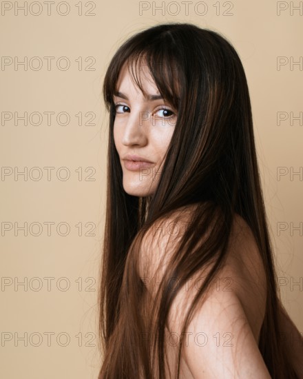 A portrait of a young woman with long brown hair, looking at camera over her shoulder on a beige