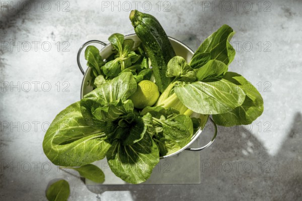 A fresh zucchini, lime, spinach and bok choy in a colander on a gray surface. The vegetables are