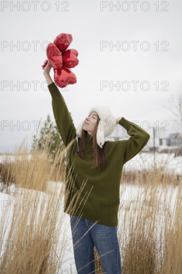 A young woman in a green sweater and white hat lifts red heart shaped balloons in a snowy park. The