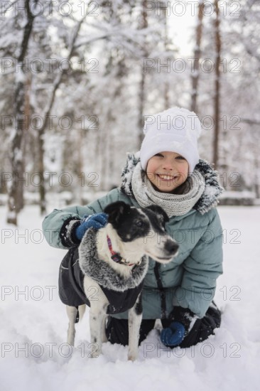 A cheerful child in winter clothing kneels in the snow with a friendly dog. Snow-covered trees
