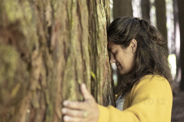 A woman in a yellow sweater hugs a tree in a forest, eyes closed, expressing a deep connection to
