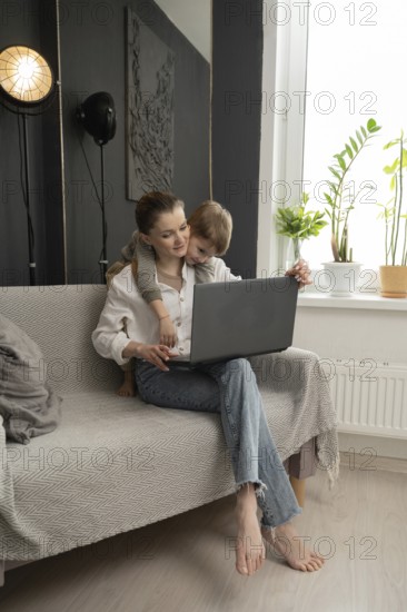A mother working on a laptop at home with her young child lovingly embracing her. A cozy, modern