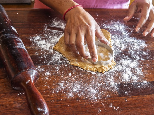 Hands using a circular cutter to shape cookie dough on a floured wooden surface. A rolling pin is