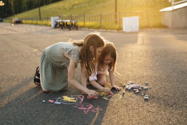 Two kids draw with colorful chalk on asphalt during a bright, sunny day, surrounded by lush
