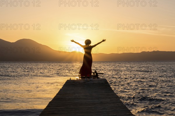 A woman stands on a wooden pier at Papikinou Beach, Milos Island, Greece, embracing the sun as it