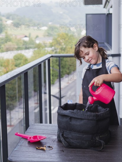 A young girl is carefully watering a sunflower in a black grow bag on a balcony. She is smiling