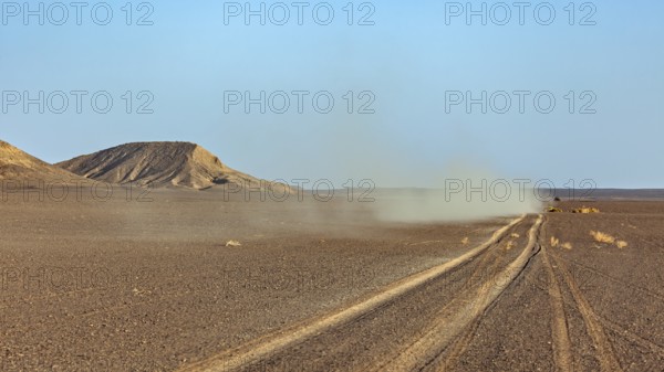 A dusty track runs through a wide, flat desert landscape with hills in the background, with an