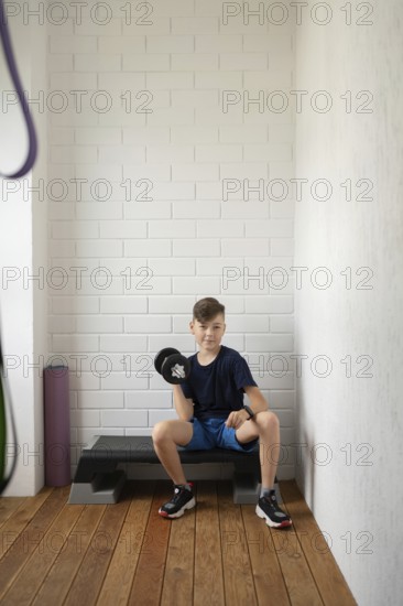 A young boy sitting on an exercise bench lifts a dumbbell in a home gym setting. He is wearing