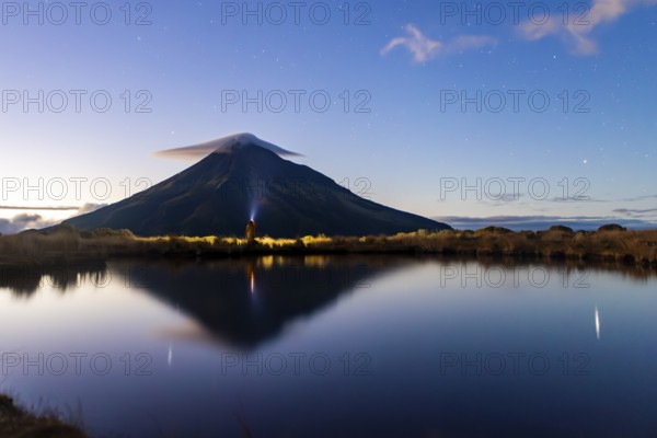Back view of unrecognizable person looking at Mount Taranaki's reflection in a tranquil lake. The