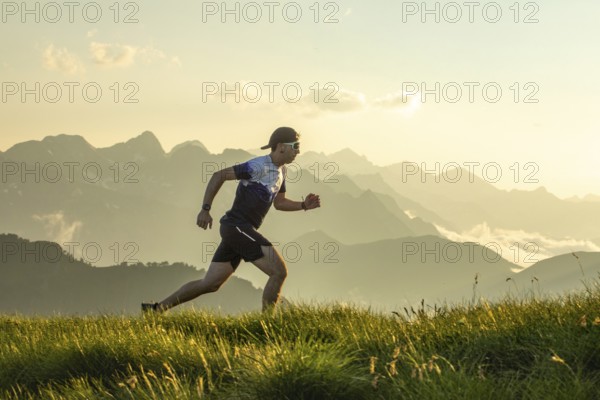 Man in athletic gear running on a grass trail with scenic mountain backdrop. The sun sets behind