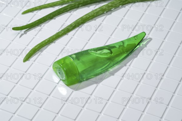 A green aloe vera gel bottle lies alongside fresh aloe leaves on a clean, white-tiled bathroom