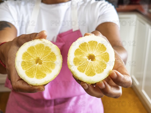 A woman in a pink apron presents two fresh lemon halves in a kitchen setting. The vivid yellow