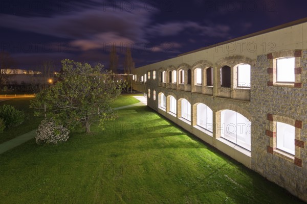 Night view of a well lit hospital with stone and brick facade. The scene includes a green lawn, a