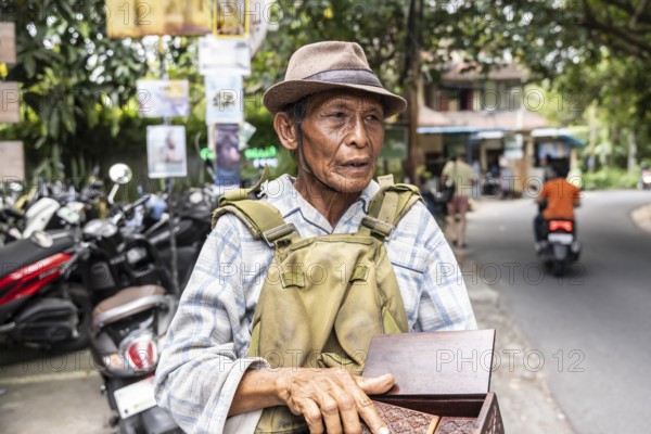 An elderly balinese man wearing a hat and plaid shirt stands on a street, holding a wooden box.