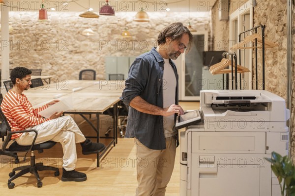 Two professionals in a cozy coworking space, one seated with documents and the other using a copier