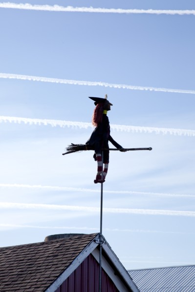 Witch and Broom on Pole Attached to Barn Roof with Airplane Trails in Sky