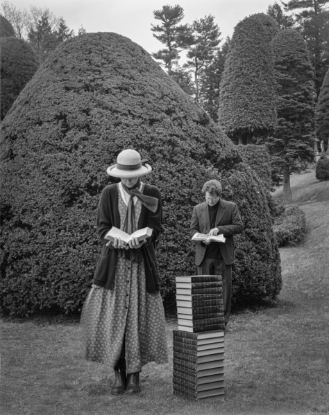 Man and woman reading while standing near stack of books in topiary garden