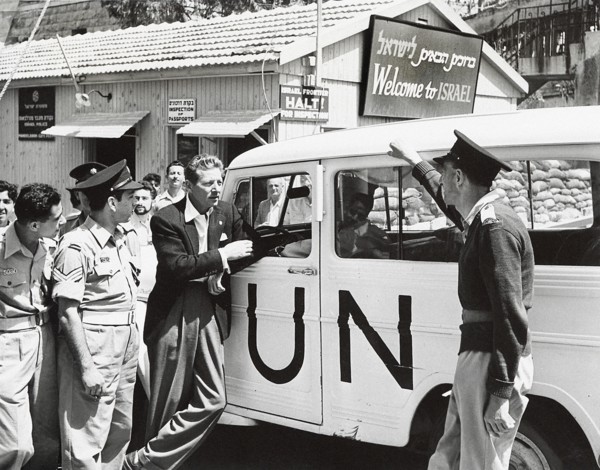 Danny Kaye leaning against UN vehicle near sign saying "Welcome to Israel"
