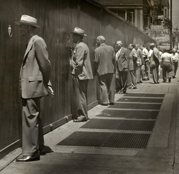Row of men standing on sidewalk looking through round peep holes at new construction site