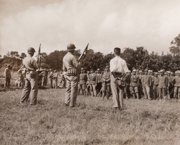 Japanese soldiers line up for formal surrender ceremonies as U.S. Marines with tommy guns and fixed