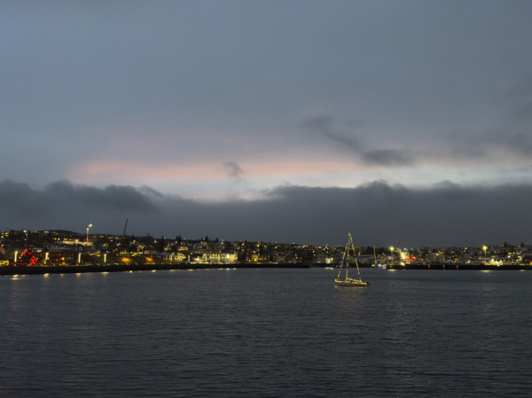 Sailboat with Christmas lights in harbor with illuminated seaside town in background,