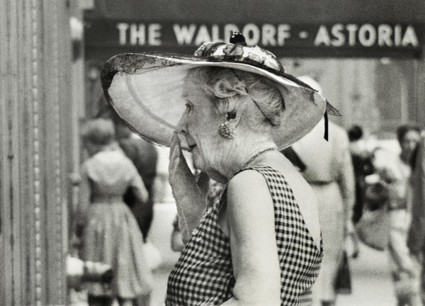 Half-length portrait of elderly woman in wide-brimmed hat standing outside Waldorf-Astoria Hotel,