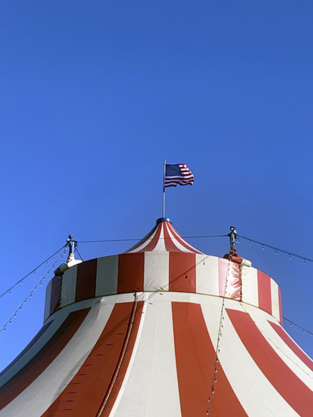 Top of red and white circus tent with American flag against blue sky