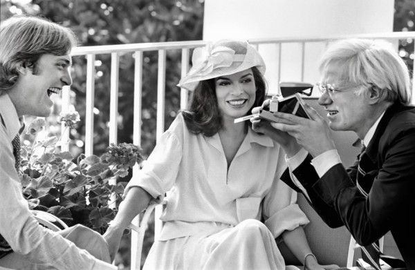 American artist Andy Warhol taking Polaroid picture while sitting with Jack Ford and Bianca Jagger