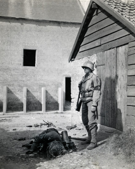 U.S. military police standing guard in barnyard near body of dead German sniper, Normandy, France,