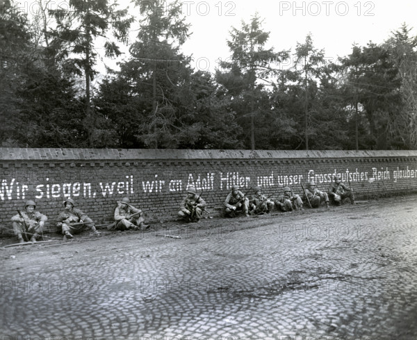 Members of 16th regiment, 1st Division, U.S. First Army, sitting under German slogan "Wir Siegen,