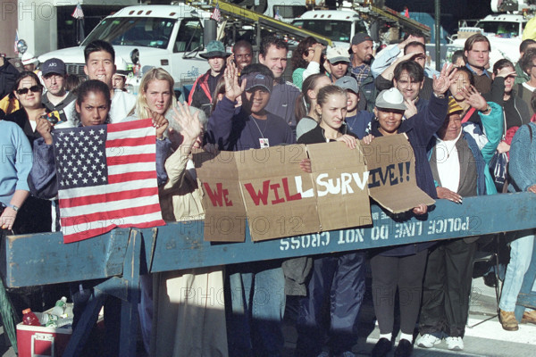 Onlookers waving and holding American flag and sign reading "We Will Survive" during U.S. President