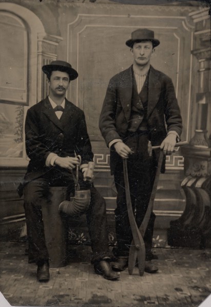 Two Stovepipe Makers, One with a Hammer and Tin Snips, the Other Sitting on Stovepipe Assembly with Tin Snips and Mallet, 1880s.