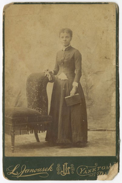Photograph of a woman standing next to a chair and holding a book, late 19th century. Creator: Louis Janousek.