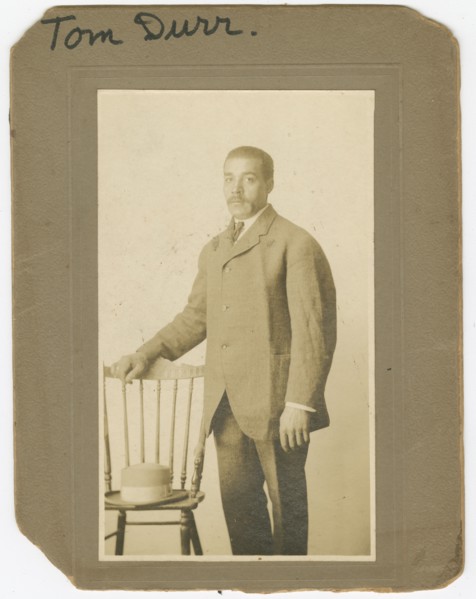 Photograph of Tom Durr standing next to a chair with a hat on it, ca. 1910. Creator: Unknown.